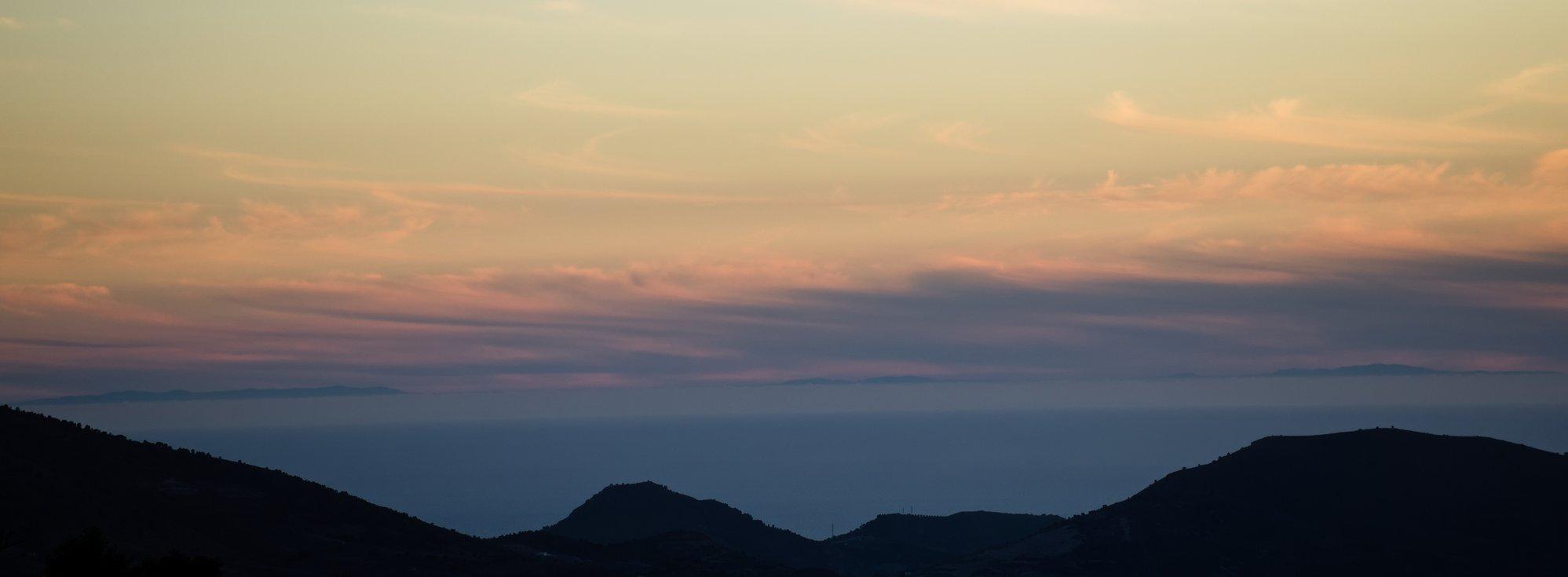 Picture at sunset looking over hills and mountains in Spain (Europe), down to the Mediterranean sea and across to the mountains on the horizon just below the changing colour of thin clouds. The mountains on the horizon are on the Rif mountains of Morocco on the continent of Africa.