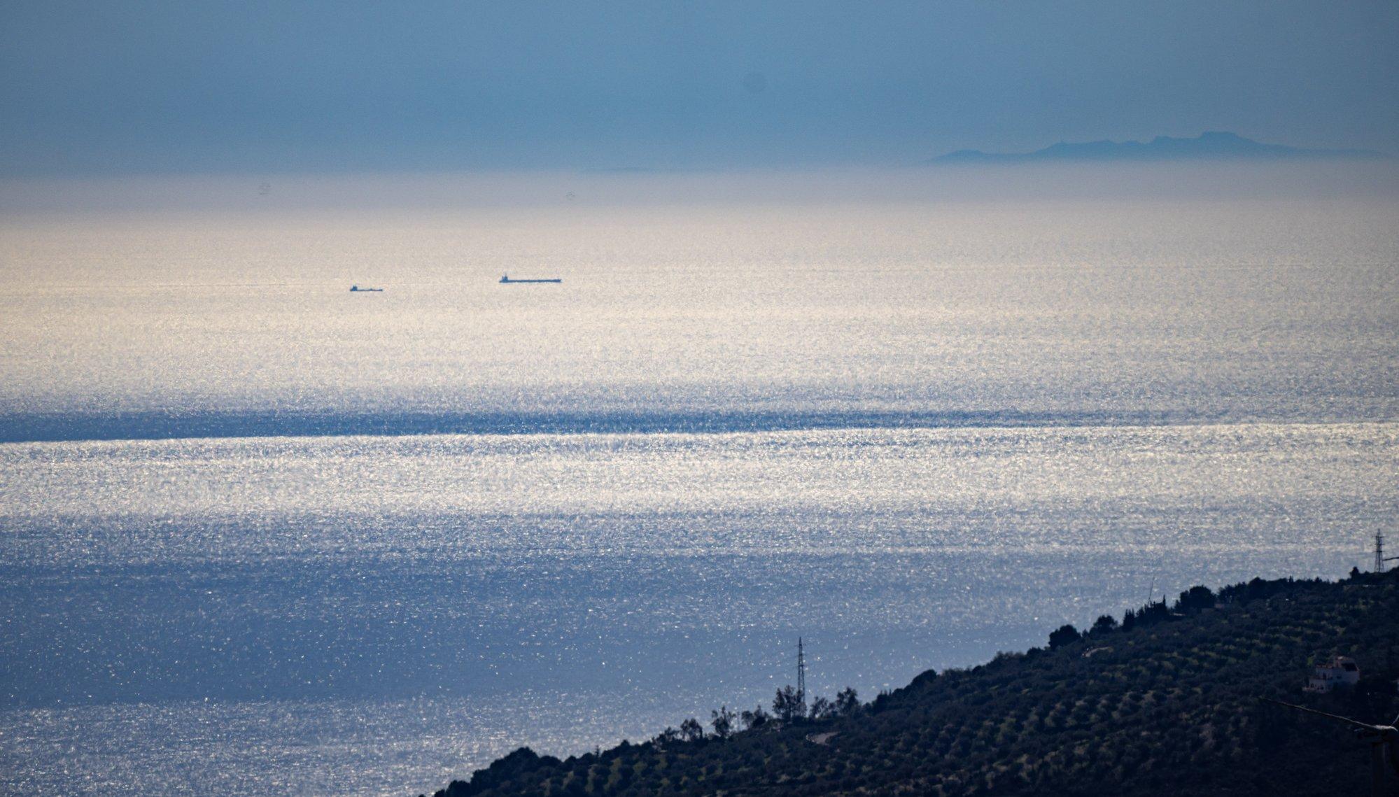 A hillside slanting down just below centre right to middle bottom. On the sunlit Mediterranean sea are two ships and behind that coming out of the haze towards the right are the Riff mountains in Morocco, Africa.