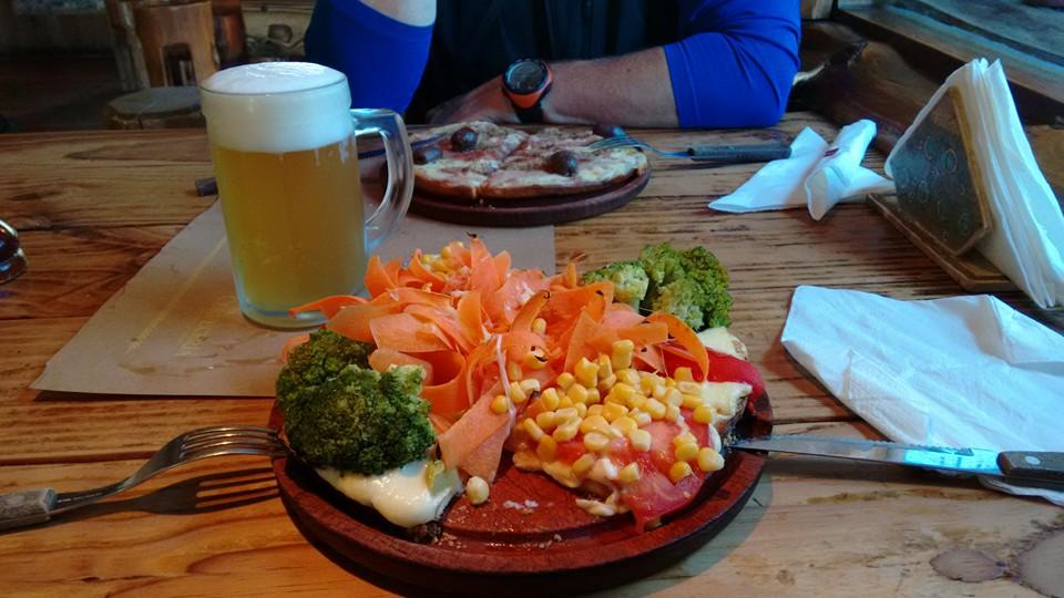 A picture showing a pizza full of vegetables. Broccoli, shaved carrot, sweetcorn are the main attraction. There is a pint of beer behind.