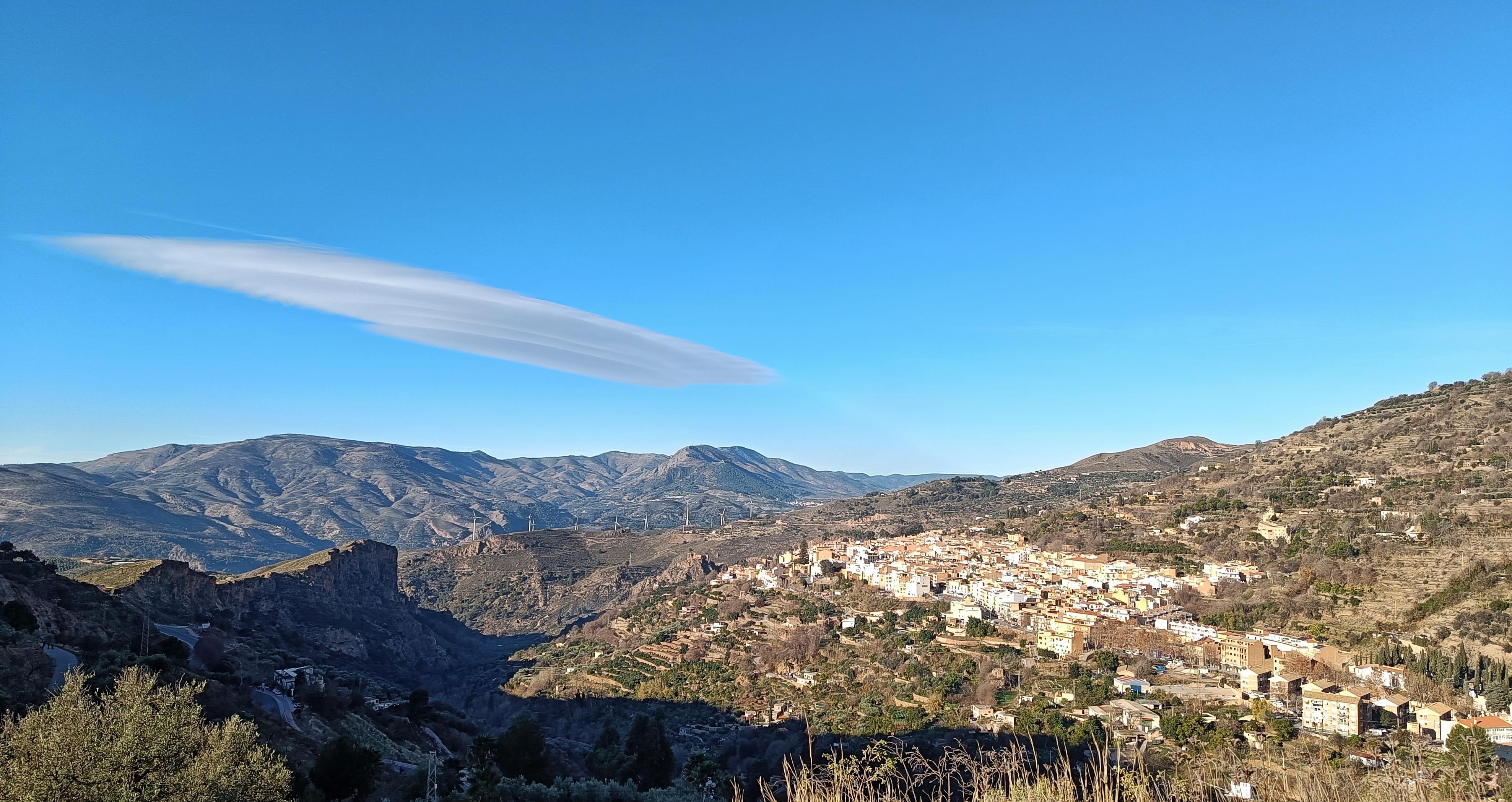 A lenticular cloud sitting in a blue sky above mountains. In the foreground a white Spanish village sits on the hillside.