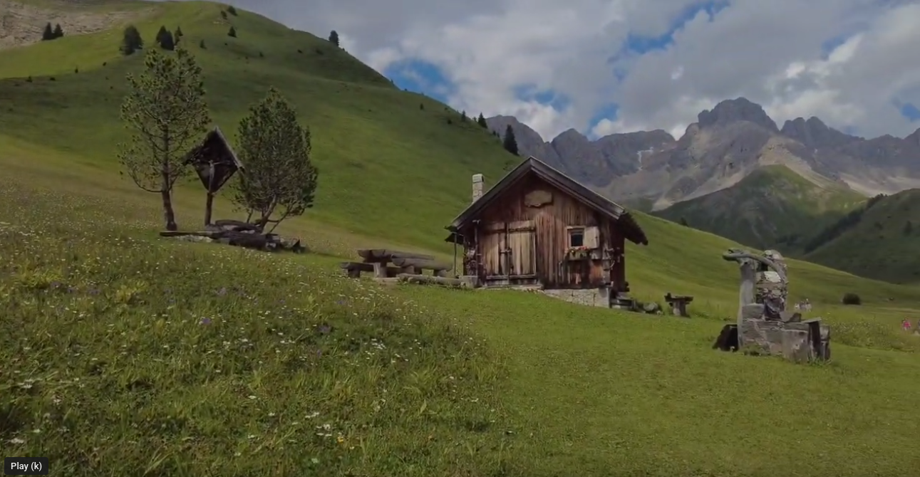 A screenshot of the video that the link in the post is about. The picture shows a wooden mountain hut in a green grass meadow. Behind the hut are high mountains, the sky has white clouds and blue showing through.