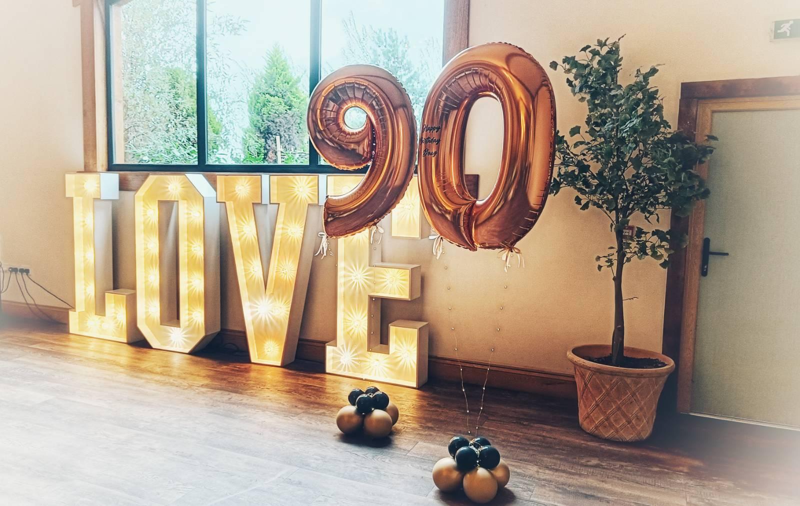 Gold balloons in the number of 90 beside a lit up sign saying love