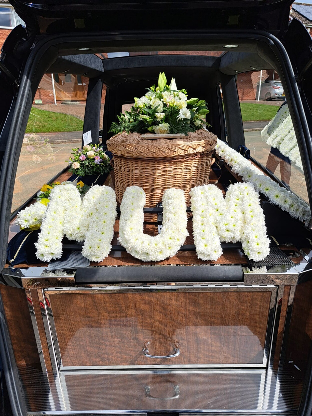 A wicker coffin inside a hearse with white flowers spelling out Mum in front.