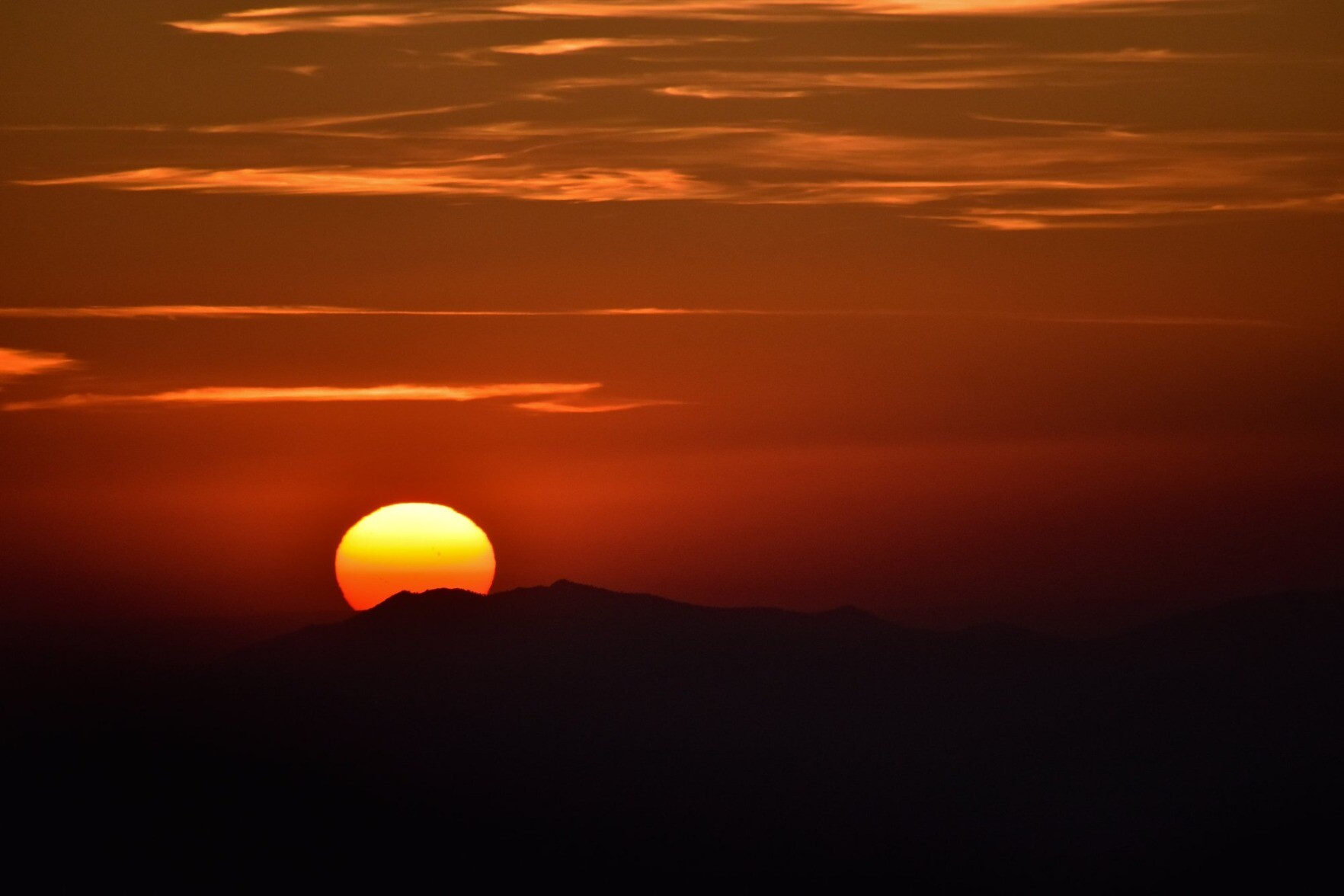 A picture of the setting sun going down behind a mountain. The mountains are silhouetted and the sky is orange