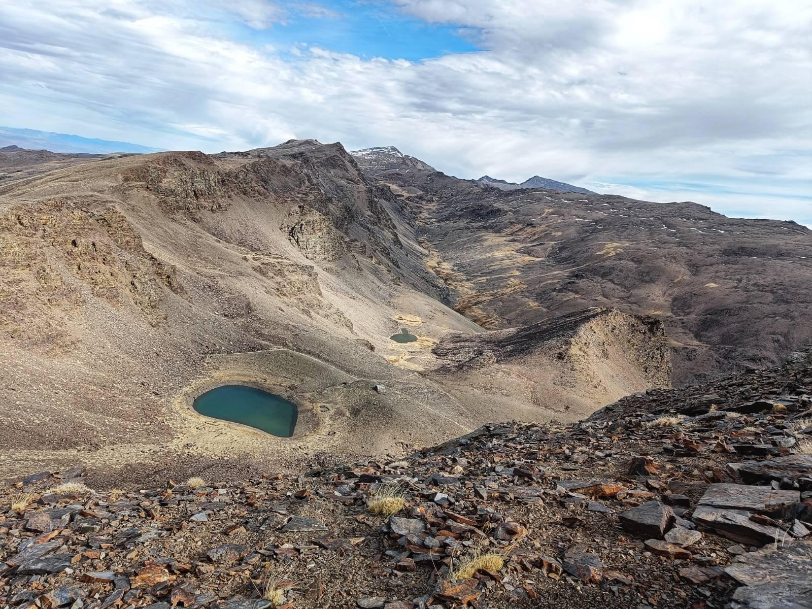 A picture showing a valley. The highest peaks of the Sierra Nevada in Spain are seen at the back of the photo with some snow clinging to them. A layer of white cloud with a hole in allowing a bit of blue sky to push through. At the bottom of the picture is a lake with a small refuge looking over it. Quite a stark image.