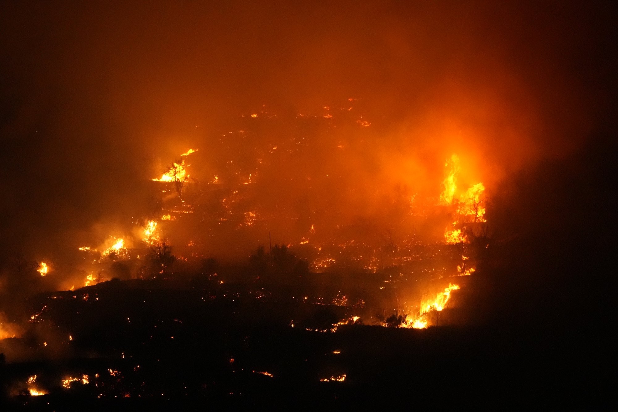 Photo of a wildfire taken at night. Orange flames,  lots of embers and smoke.