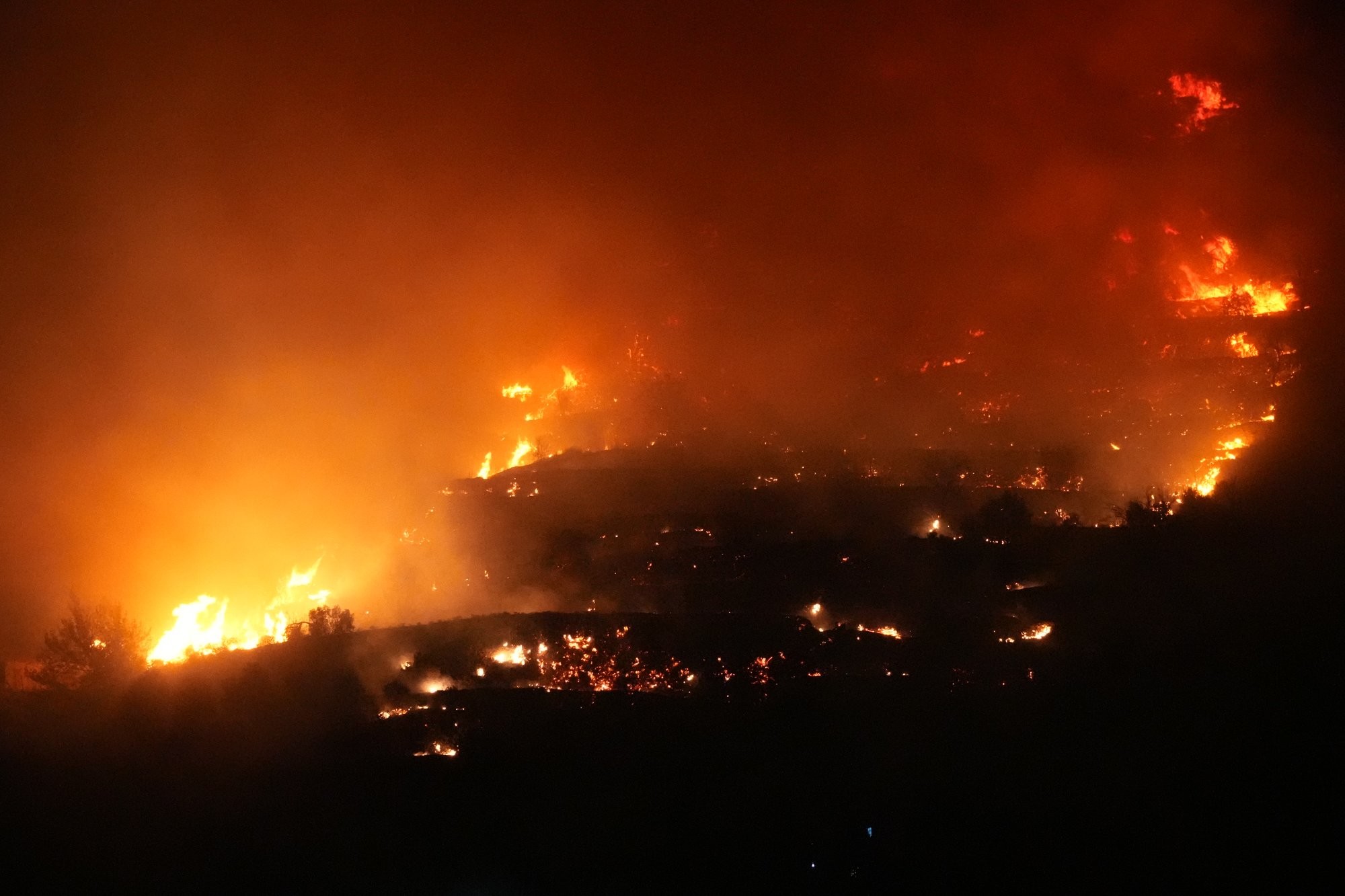 Photo of a wildfire taken at night, yellow flames on the left and orange flames at the top and right. 