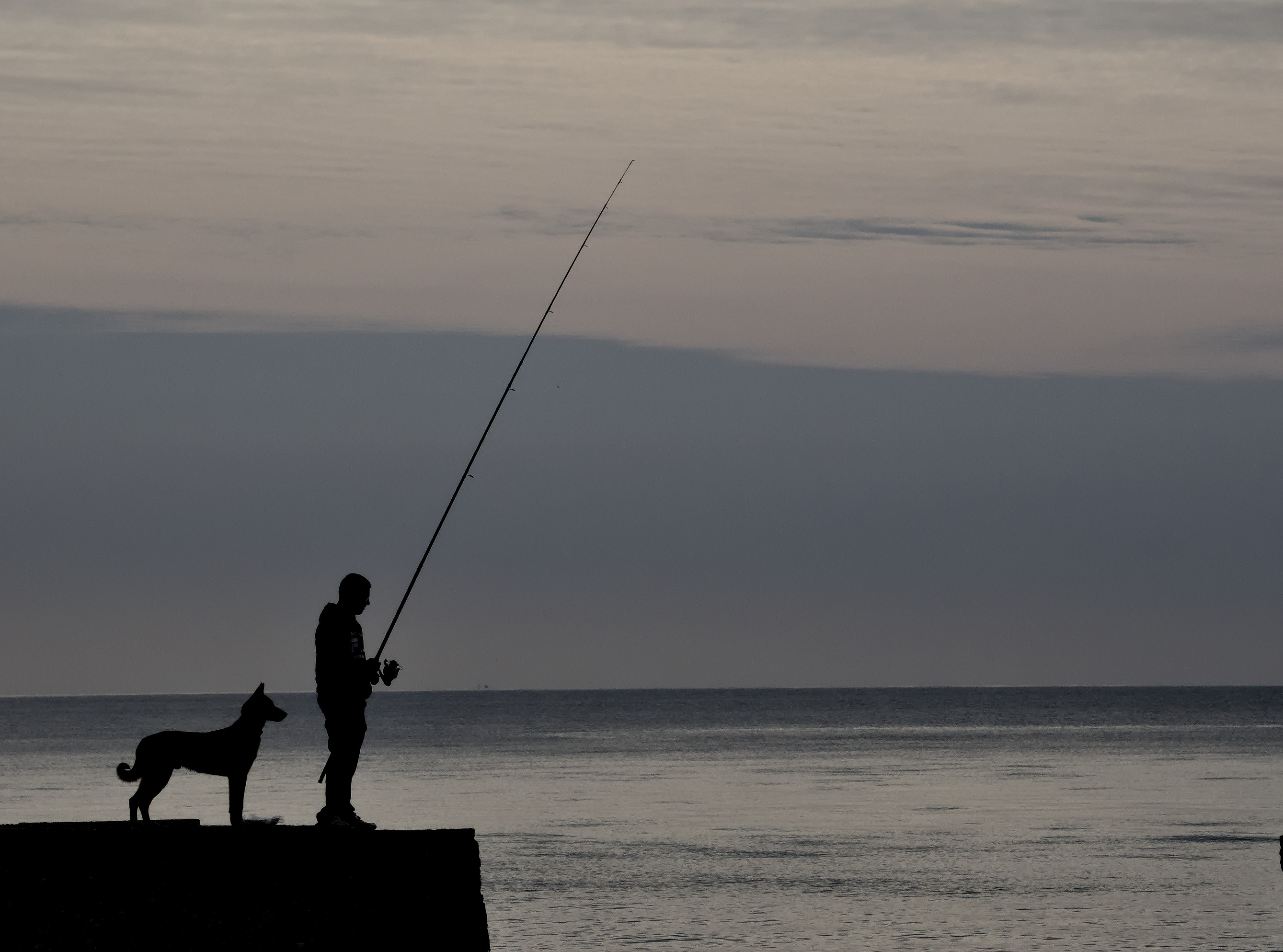 The almost silhouette of a dog stood behind it's owner who is holding a long fishing rod and is fishing off the end of a wall. The calm grey Mediterranean sea and grey sky.