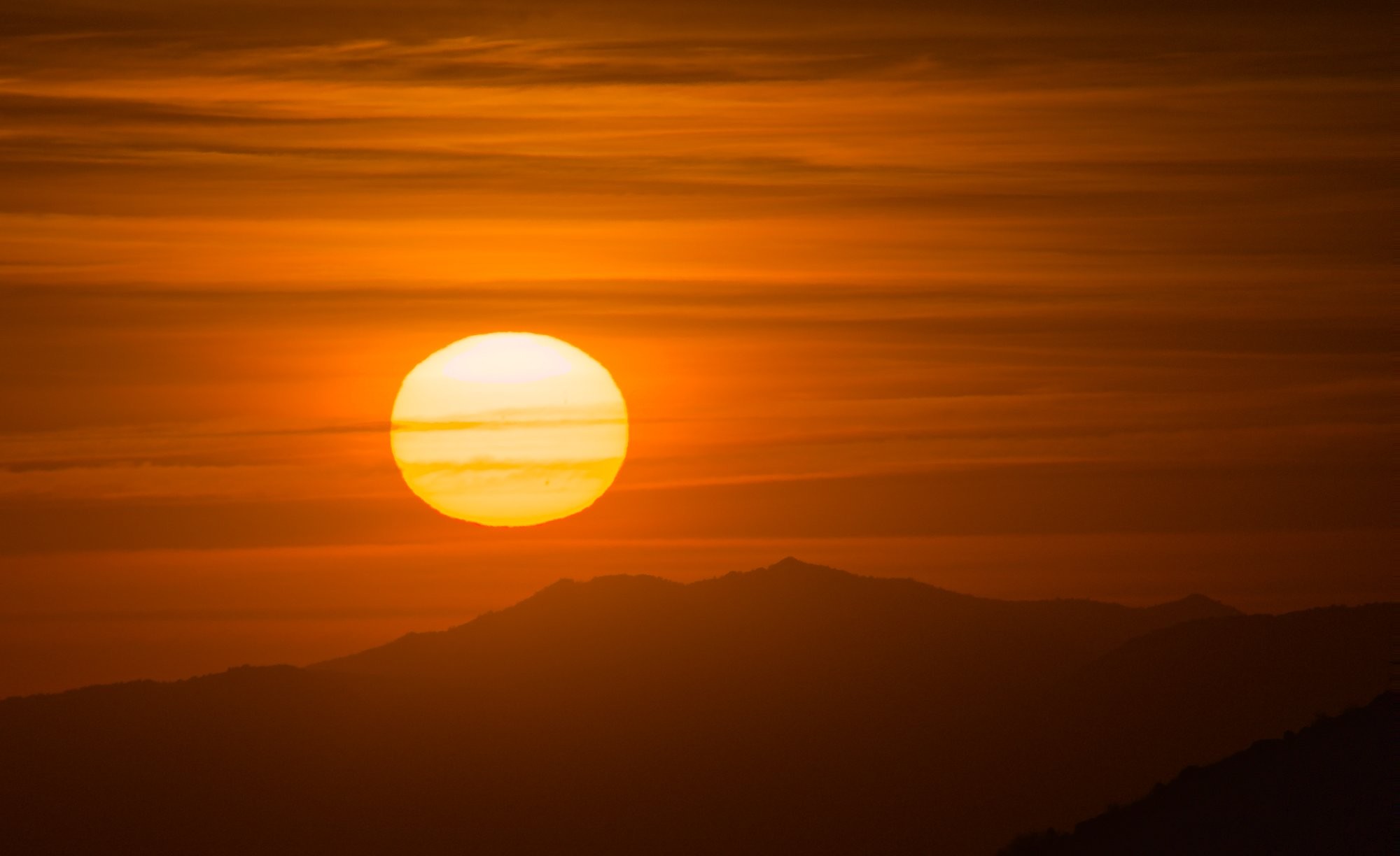 The setting sun just above some mountain ridges in the distance with the light clouds and mountains turned orange