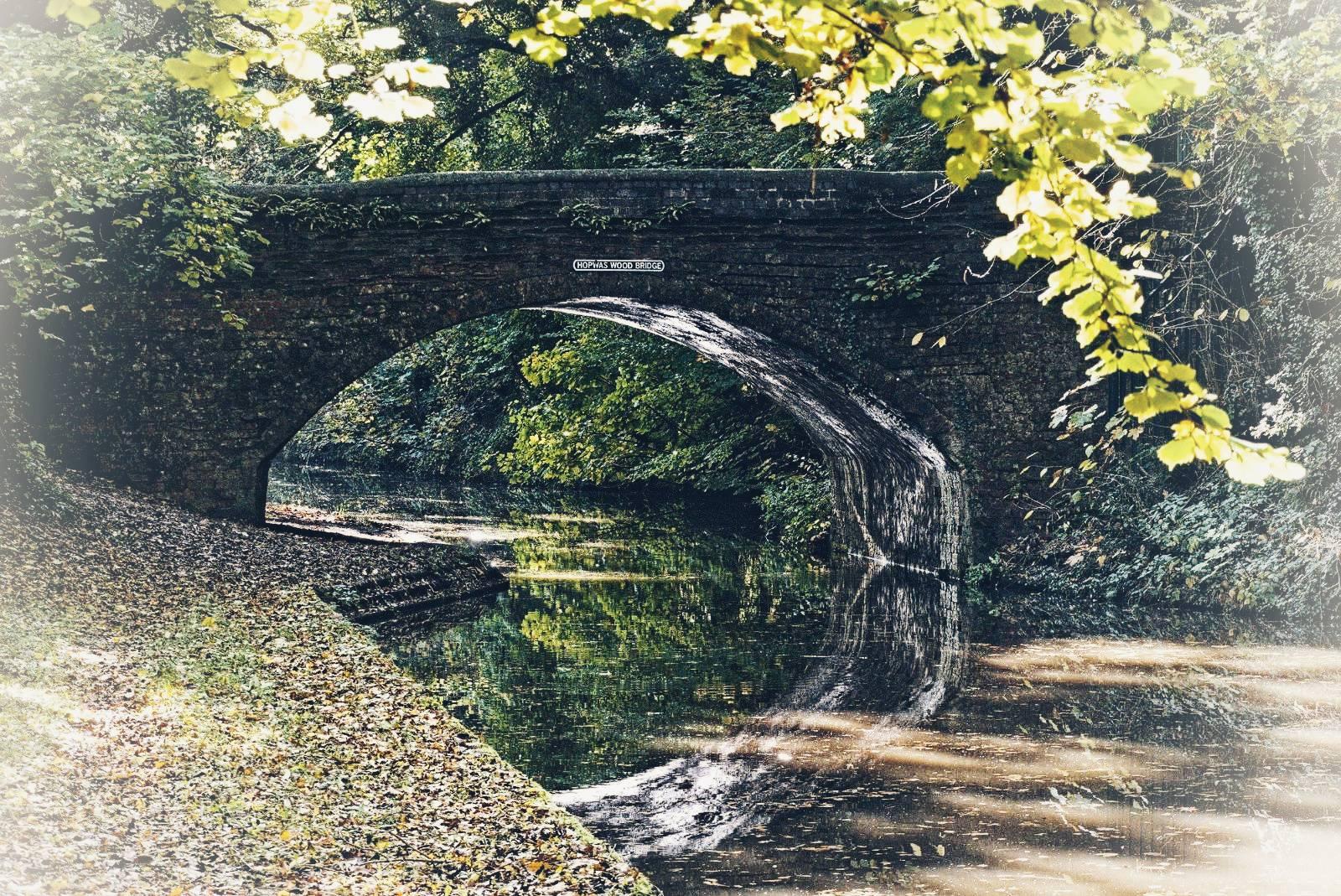 A canal bridge with it's reflection in the canal. Autumn colours just starting on the trees which are also reflected in the water, fallen leaves on the tow path.