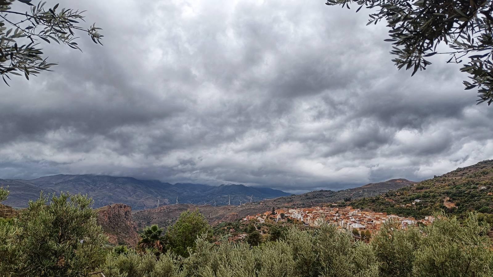 A Spanish village on a hillside surrounded by trees, mountains, wind turbines all looking very wet underneath a various shades of grey coloured sky.