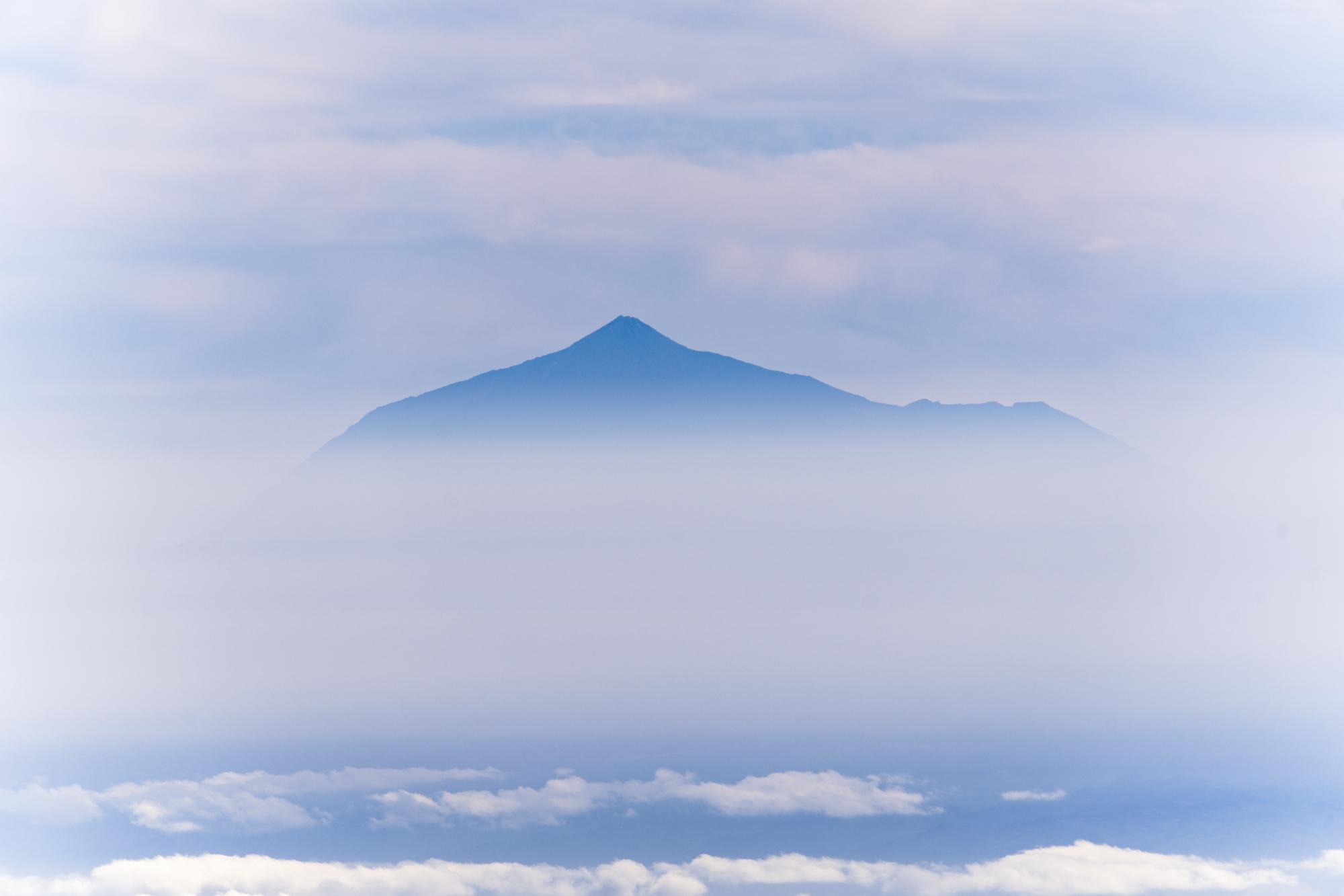 A misty picture of a volcano coming out of the mist. Clouds above and clouds over the see below.