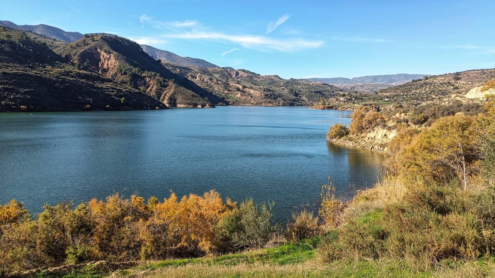 A blue lake surrounded by hillsides covered in trees and shrubs. In for foreground a bit of grass and some autumn coloured shrubs.